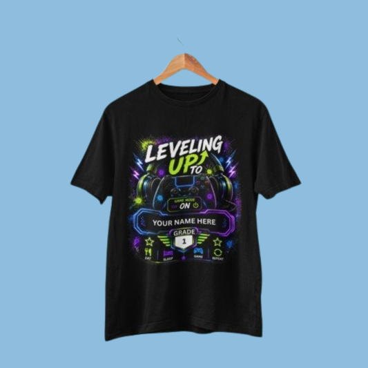A black short-sleeve t-shirt hanging on a wooden hanger against a solid light blue background. The shirt features a colorful graphic print with gaming-themed text reading 'LEVELING UP TO YOUR NAME HERE' and 'GRADE 1' on a black banner.