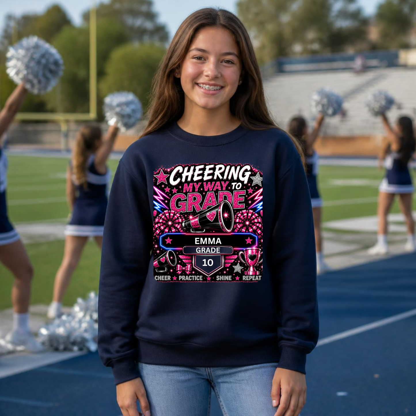 A smiling young woman stands on a school track field wearing a navy blue crewneck sweatshirt with a colorful graphic that reads 'CHEERING MY WAY TO GRADE' and 'EMMA GRADE 10'. Cheerleaders are visible in the blurred background.