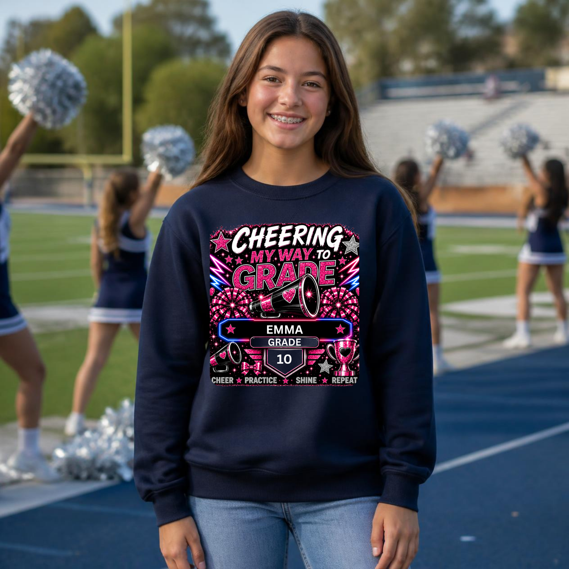 A smiling young woman stands on a school track field wearing a navy blue crewneck sweatshirt with a colorful graphic that reads 'CHEERING MY WAY TO GRADE' and 'EMMA GRADE 10'. Cheerleaders are visible in the blurred background.