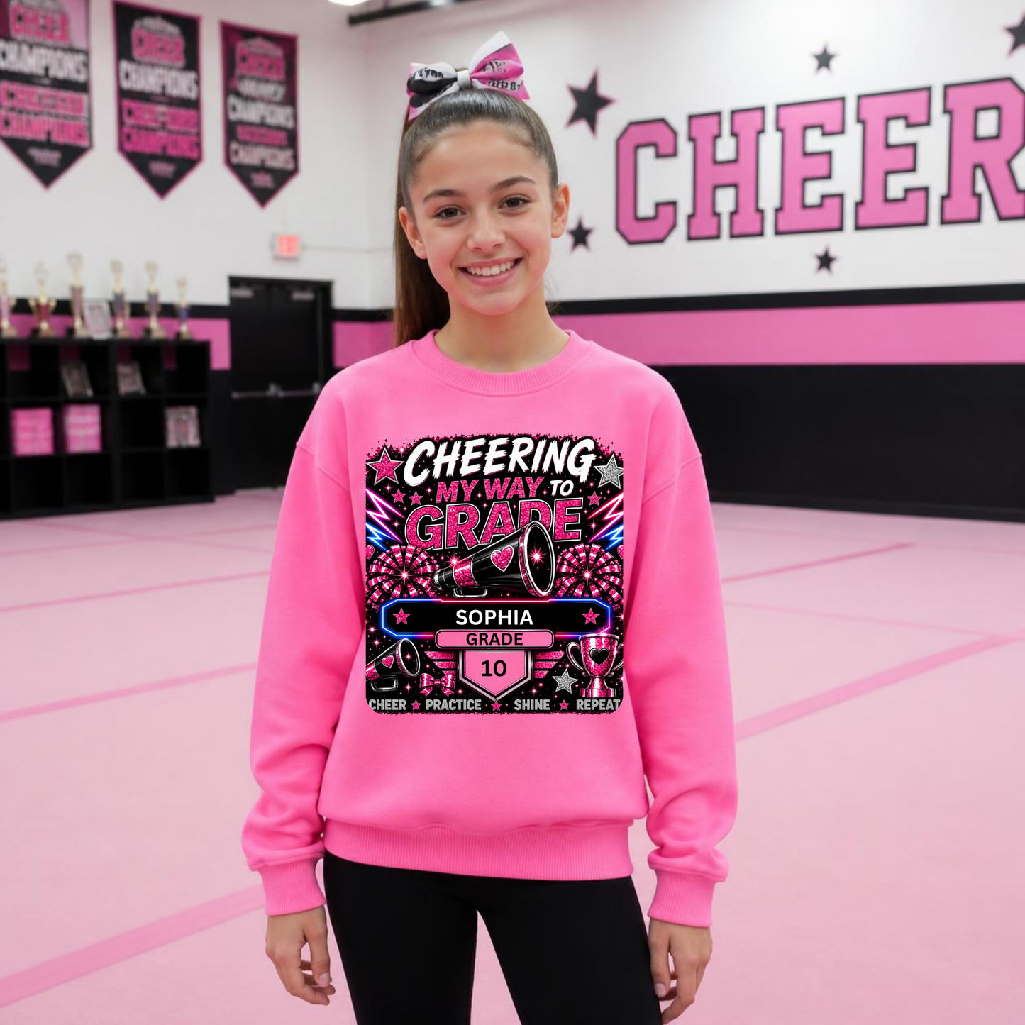 A young girl with a pink hair bow stands in a cheerleading gym wearing a bright  safety pink sweatshirt with a custom graphic that reads 'CHEERING MY WAY TO GRADE' and 'SOPHIA GRADE 10'. The background features a pink and white wall with 'CHEER' written on it.