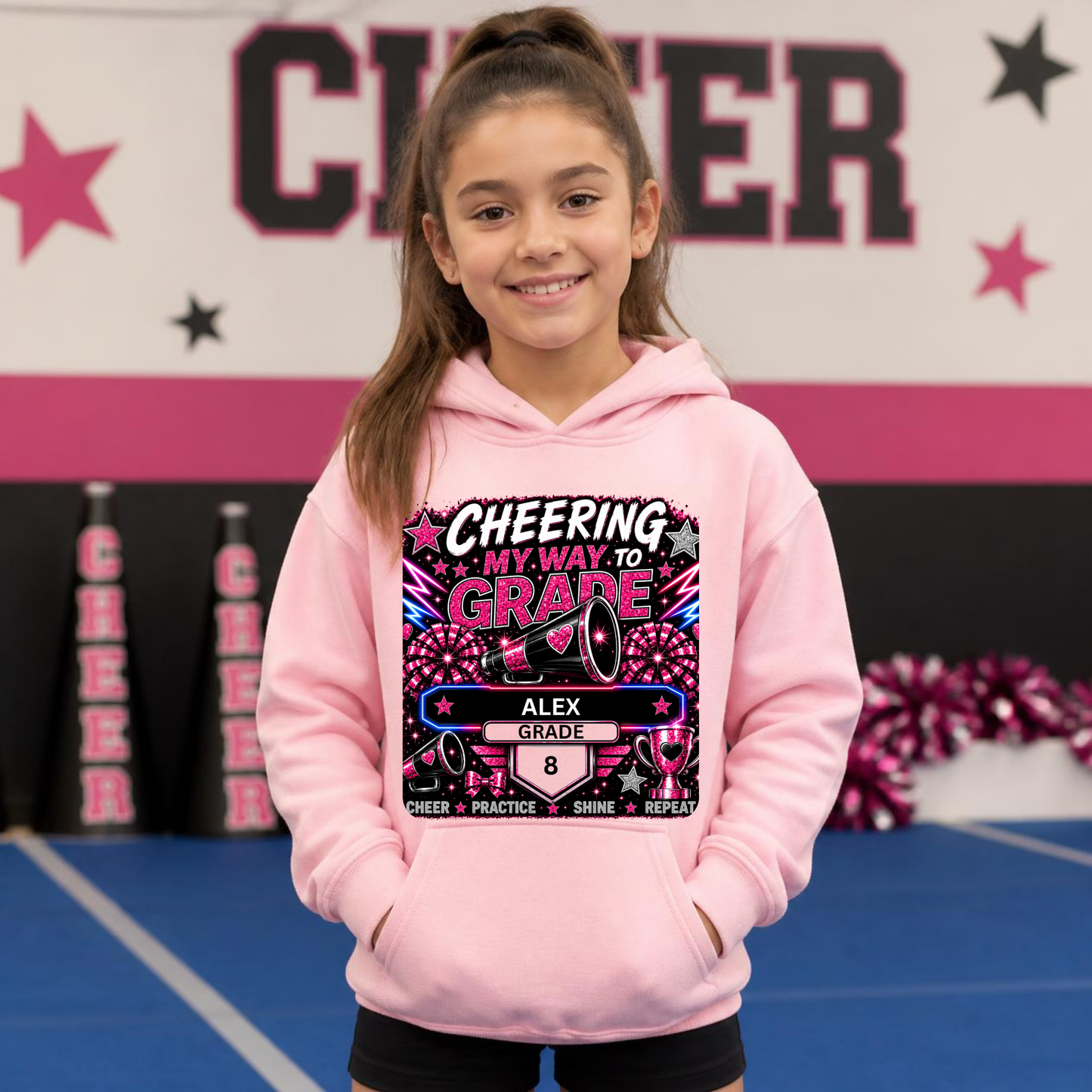 A young girl with a ponytail stands on a cheerleading mat wearing a light pink hoodie with a colorful graphic that reads 'CHEERING MY WAY TO GRADE ALEX GRADE 8'.