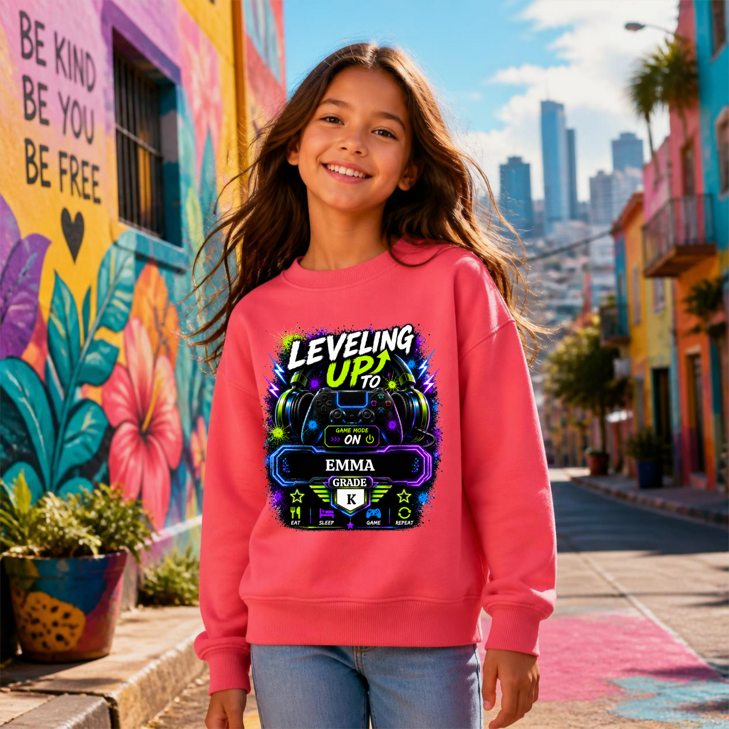 A smiling young girl stands on a colorful city street wearing a bright pink crewneck sweatshirt with a large graphic print that reads 'LEVELING UP TO EMMA GRADE K'. The background features vibrant, painted murals on the left wall and a sunny urban street scene with buildings under a blue sky.