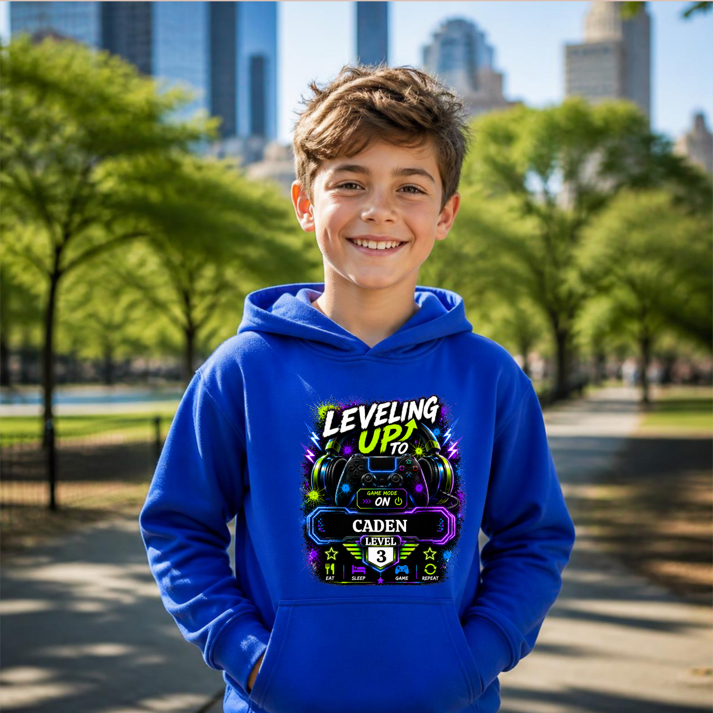 A smiling young boy wearing a bright blue hoodie with a colorful video game graphic on the chest, standing in a sunny park with trees and city buildings blurred in the background.