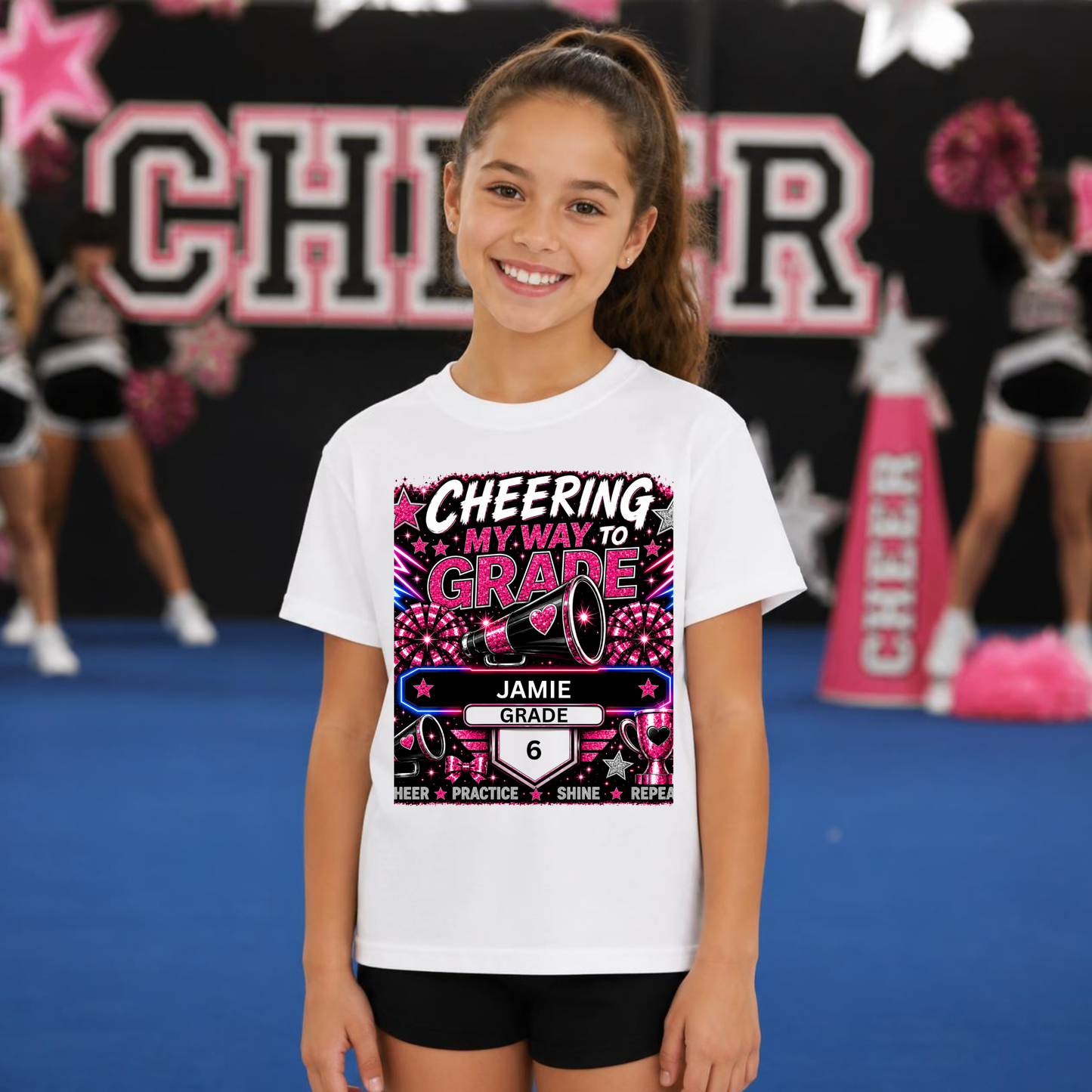 A young girl smiling on a blue cheerleading mat, wearing a white graphic t-shirt with 'Cheering My Way To Grade 6' printed in a colorful design. Cheerleaders are visible in the blurred background.