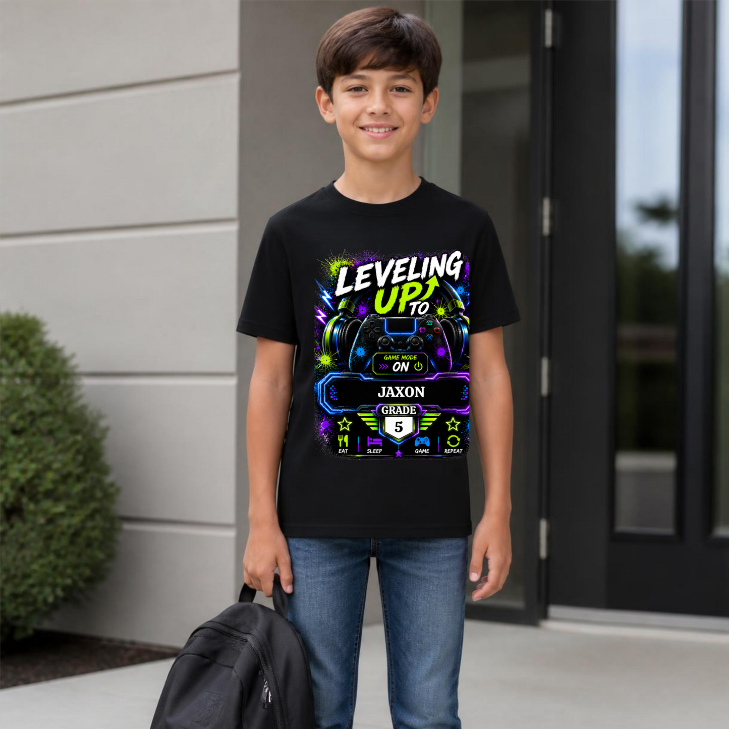 A young boy wearing a black graphic t-shirt with a neon gaming theme that reads 'LEVELING UP! TO JAXON GRADE 5'. He is standing outdoors in front of a modern house wall and glass doors, holding a black backpack.