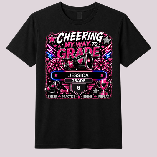 A black short-sleeve crewneck t-shirt featuring a colorful graphic print on the front. The graphic displays a megaphone, fireworks, and trophies with the text 'CHEERING MY WAY TO GRADE', 'JESSICA', 'GRADE 6', and 'CHEER PRACTICE SHINE REPEAT' in vibrant pink, purple, and white colors.
