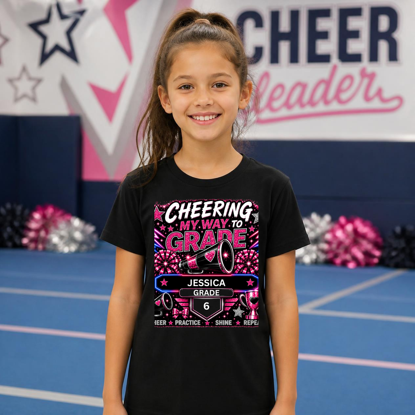 A young girl with her hair in a high ponytail stands in a cheerleading gym wearing a black graphic t-shirt. The shirt features a colorful design with the text 'CHEERING MY WAY TO GRADE' and a mock grading badge for 'JESSICA GRADE 6'.
